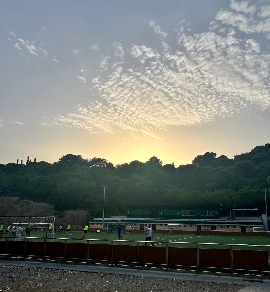A floodlit five-a-side pitch below a wooded hillside at dusk, players in yellow bibs mid-game under a mackerel sky.