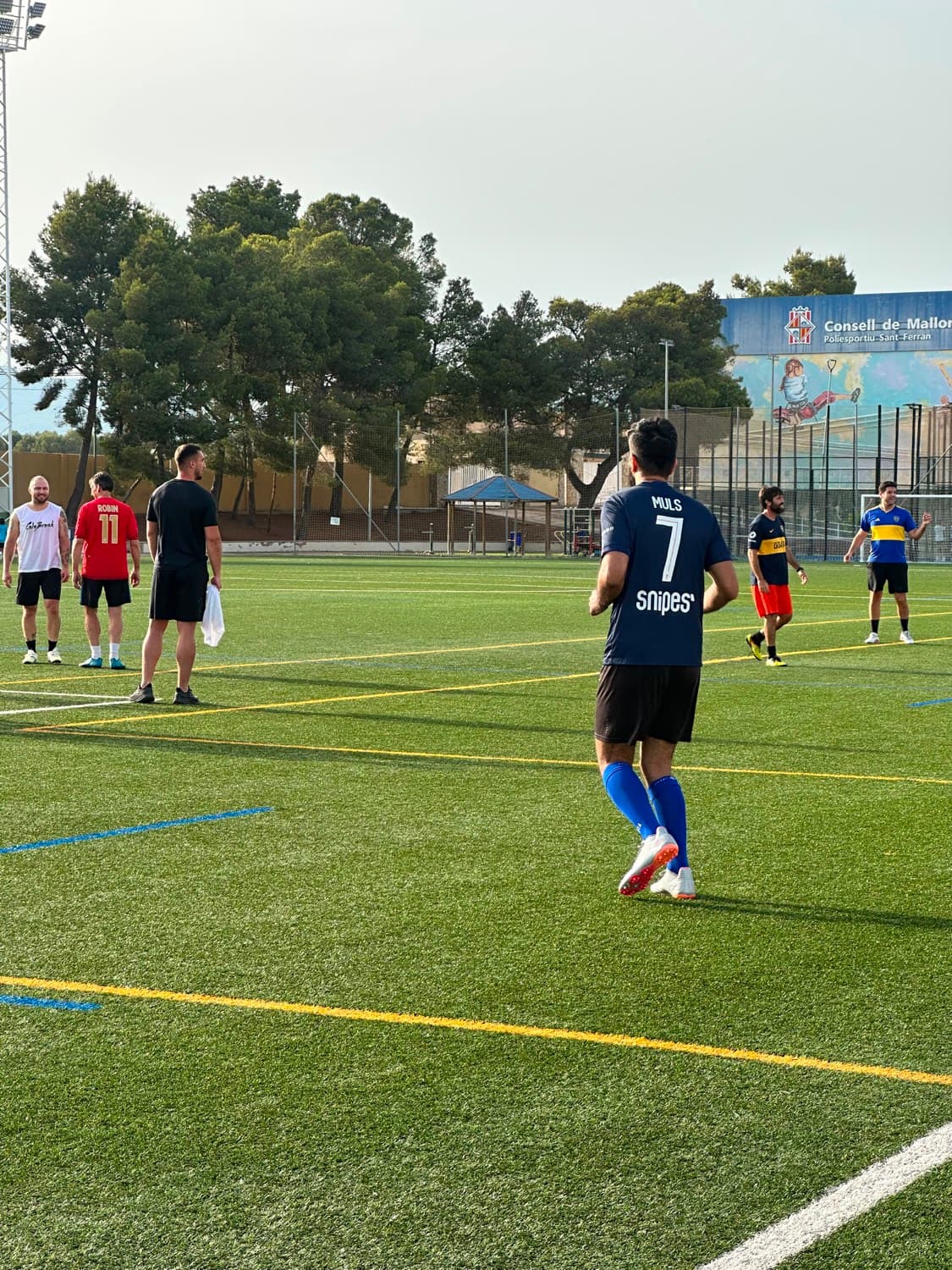 A pickup game warming up on a green artificial pitch, the writer walking away from camera in a number 7 shirt.