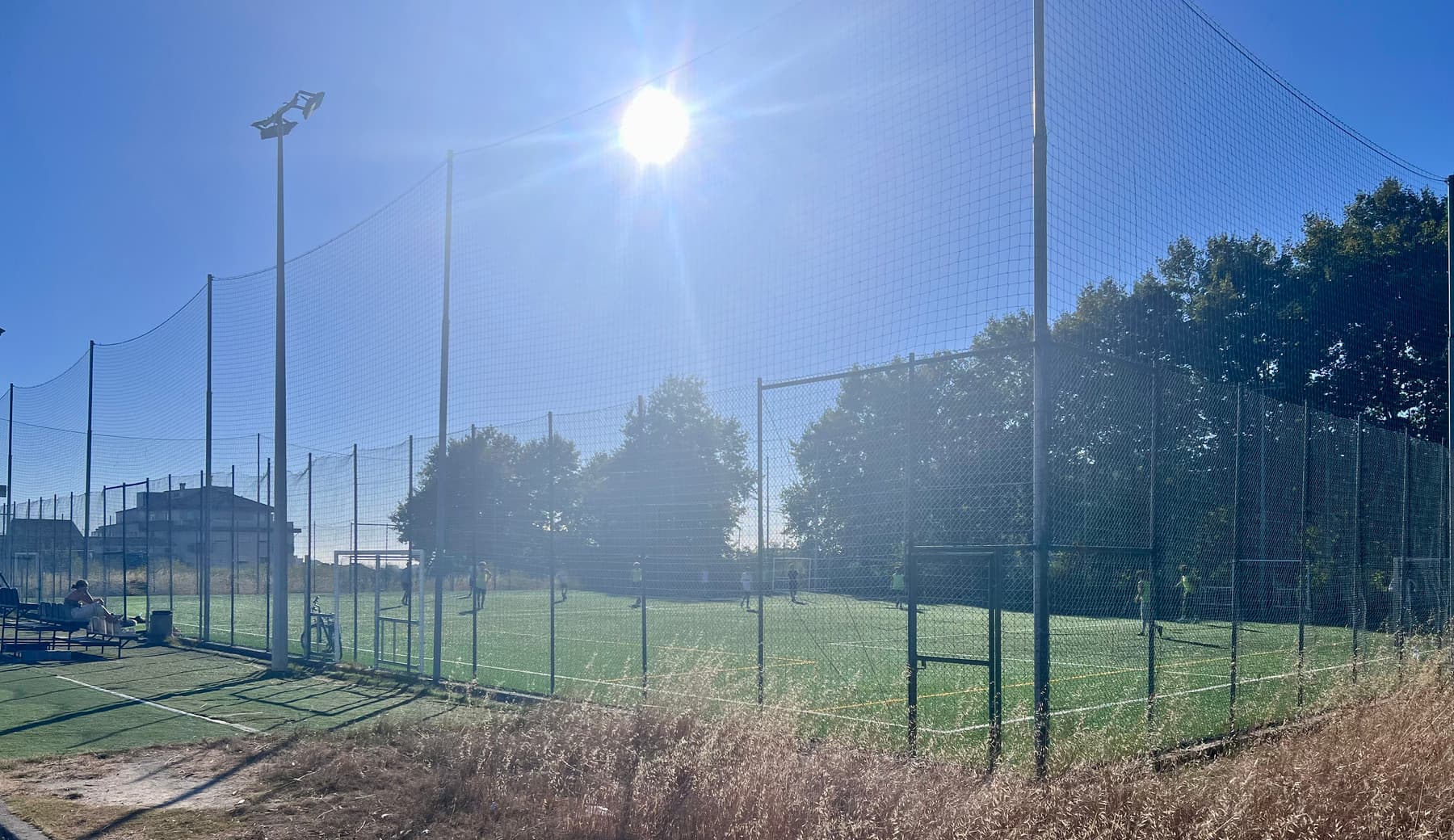 Two caged artificial pitches in hard midday sun, seen through tall ball-stop netting from a path of dry grass.